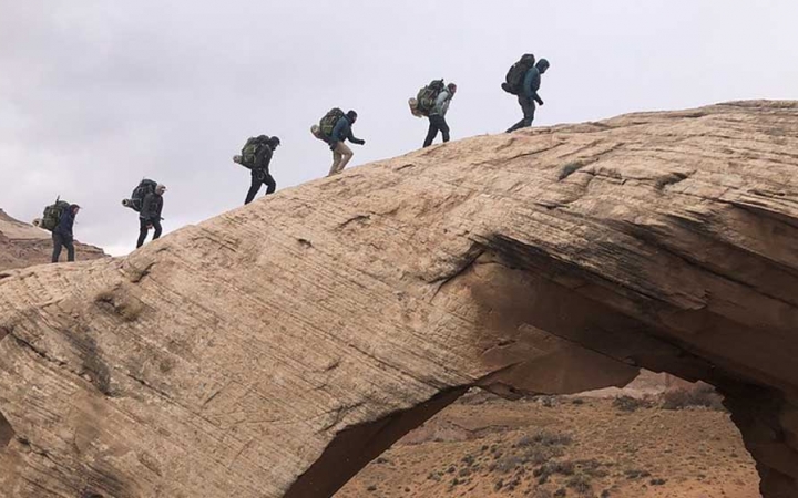 a group of gap year students hike in a line over a rock arch in the american southwest